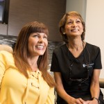 Two women in dental office consultation - patient in yellow top seated in treatment chair speaking with staff member in black scrubs